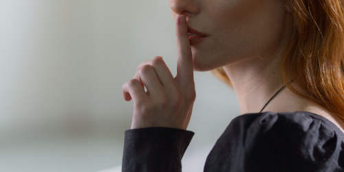 A woman is pressing her finger to her mouth as a gesture to request silence.